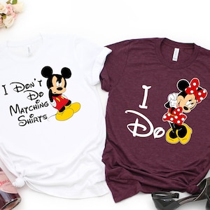 Op de afbeelding: Twee witte T-shirts met een Mickey Mouse en Minnie Mouse ontwerp. Het witte shirt zegt "I Don't Do Matching Shirts" en toont Mickey Mouse met een chagrijnige uitdrukking. Het bordeauxrode shirt zegt "I Do" en toont Minnie Mouse met een gelukkige uitdrukking.