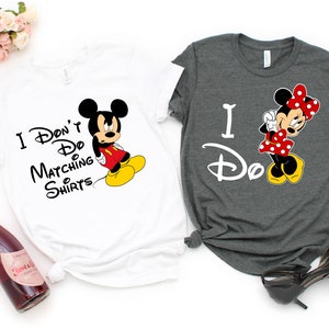 Op de afbeelding: Twee T-shirts met een Mickey Mouse en Minnie Mouse ontwerp. Het witte T-shirt zegt "I Don't Do Matching Shirts" en het grijze T-shirt zegt "I Do".