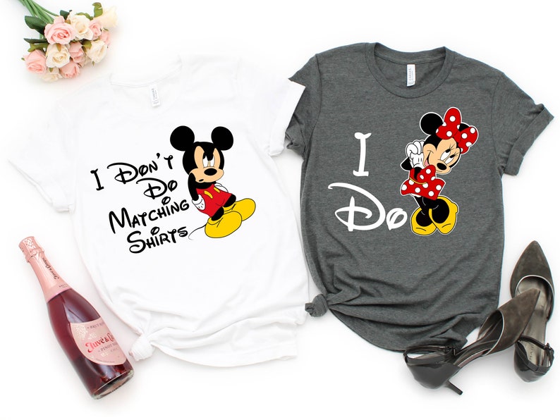 Op de afbeelding: Twee T-shirts met een Mickey Mouse en Minnie Mouse ontwerp. Het witte T-shirt zegt "I Don't Do Matching Shirts" en het grijze T-shirt zegt "I Do".