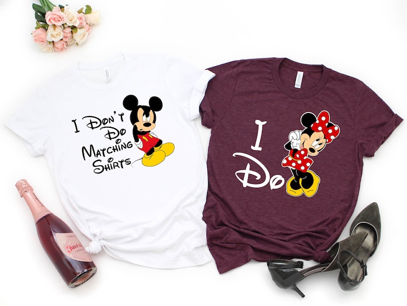 Op de afbeelding: Twee witte T-shirts met een Mickey Mouse en Minnie Mouse ontwerp. Het witte shirt zegt "I Don't Do Matching Shirts" en toont Mickey Mouse met een chagrijnige uitdrukking. Het bordeauxrode shirt zegt "I Do" en toont Minnie Mouse met een gelukkige uitdrukking.