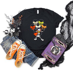 May include: Black t-shirt with a cartoon image of Donald Duck dressed as a skeleton holding a bunch of Mickey Mouse head balloons with jack-o-lantern faces. The balloons are orange, yellow, and green. The shirt has a crew neck and short sleeves.