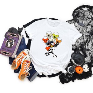 May include: White t-shirt with a cartoon skeleton wearing a cape and holding orange, yellow, and green balloons with Mickey Mouse ears. The balloons have spooky faces.