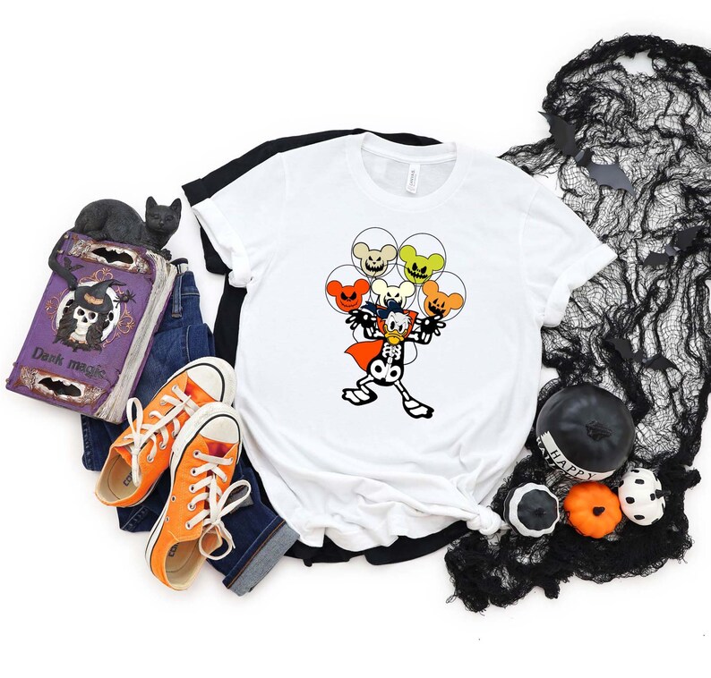 May include: White t-shirt with a cartoon skeleton wearing a cape and holding orange, yellow, and green balloons with Mickey Mouse ears. The balloons have spooky faces.