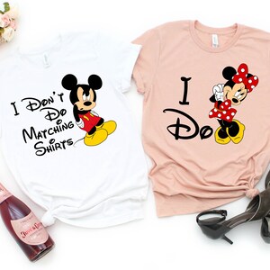 Op de afbeelding: Twee witte T-shirts met een Mickey Mouse en Minnie Mouse ontwerp. Het Mickey Mouse shirt zegt "Ik doe geen matchende shirts". Het Minnie Mouse shirt zegt "Ik doe het".