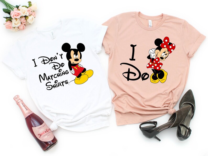 Op de afbeelding: Twee witte T-shirts met een Mickey Mouse en Minnie Mouse ontwerp. Het Mickey Mouse shirt zegt "Ik doe geen matchende shirts". Het Minnie Mouse shirt zegt "Ik doe het".