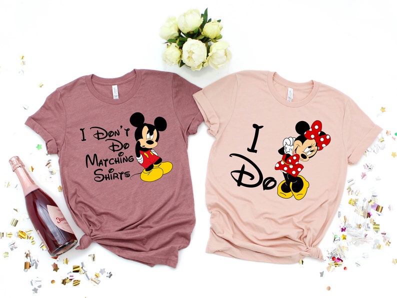 Op de afbeelding: Twee T-shirts met een Disney-thema. Het roze shirt zegt "Ik doe het" met een afbeelding van Minnie Mouse. Het mauve shirt zegt "Ik doe geen matchende shirts" met een afbeelding van Mickey Mouse.