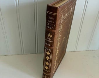 Puede incluir: Un libro encuadernado en cuero marrón titulado The Call of the Wild de Jack London. El lomo presenta letras doradas y motivos de hojas decorativos. La portada muestra un borde grabado en oro y los bordes de las páginas están dorados.