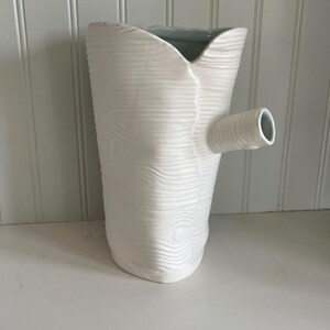 May include: White ceramic vase with a textured wood grain design and a spout. The vase has a light blue glaze on the inside.