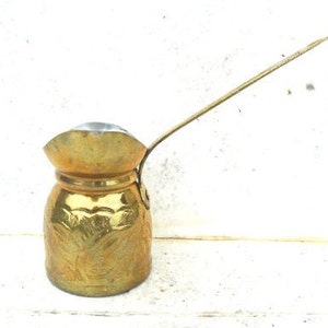Peut inclure: Une cafetière en métal doré avec une longue poignée et un design orné. La cafetière a un corps rond et un bec verseur sur le côté.