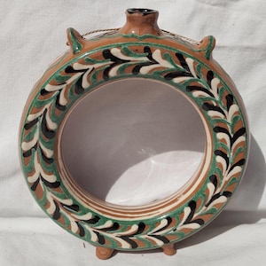 Op de afbeelding: Een ronde keramische fles met een decoratief bloemmotief in groen, zwart en wit op een bruine achtergrond. De fles heeft een cirkelvormige opening en kleine voetjes. Bovenop zit een klein handvat.
