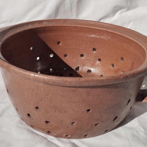 Może przedstawiać: Brązowa ceramiczna durszlak z uchwytem. Durszlak ma okrągły kształt z licznymi małymi otworami w całej misce. Uchwyt jest zakrzywiony i przymocowany do boku miski. Durszlak stoi na białej powierzchni.