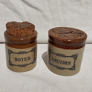 May include: Two small, vintage ceramic jars with cork lids. One jar is labelled "Boter" and the other "Kruiden" in black text. The jars are tan with brown accents and decorative label borders. The jars are likely for storing food items.