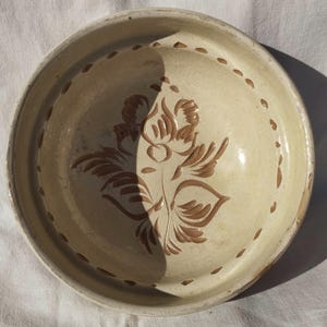 May include: Cream-colored ceramic bowl with a floral design in brown. The bowl has a wide rim with dotted accents and a glossy finish. The floral pattern is centered in the bowl.