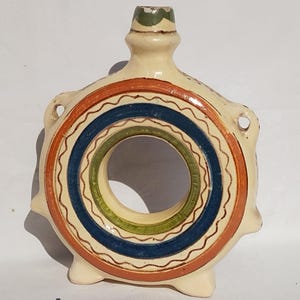May include: A white ceramic bottle with a ring-shaped opening and colourful painted designs. The bottle has a narrow neck and a small spout. The design features concentric circles in shades of blue, green, and orange.