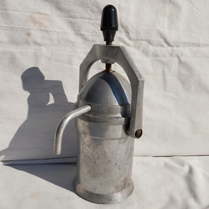 May include: Vintage manual espresso maker with a silver-colored body, a curved spout, and a black handle. The espresso maker has a cylindrical base and a domed top with a handle. The overall design is industrial and functional.