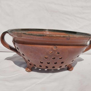 May include: A brown glazed ceramic colander featuring two side handles and three small feet. The bowl has a wide rim and a series of circular drainage holes arranged in a triangular pattern on the sides and bottom of the vessel.