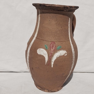 Op de afbeelding: Een bruine terracotta kan met een brede rand en een handvat. De buitenkant is voorzien van verticale witte strepen en een centraal bloemmotief in wit, groen en rood. Het oppervlak heeft een matte, getextureerde afwerking.