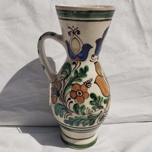 Puede incluir: Una jarra de cerámica de color crema con un motivo floral pintado a mano en tonos azul, naranja y verde. El recipiente tiene un borde acampanado, un asa curva y un cuerpo redondeado que se estrecha hacia una base circular con bandas verdes.