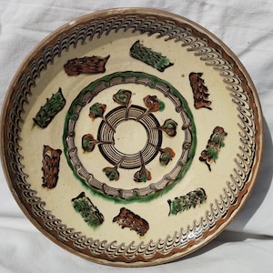 May include: A round, cream-colored ceramic plate with a brown and green decorative pattern. The plate features a central circular design and repeating leaf-like shapes around the rim. The plate's edge is trimmed with a brown border.