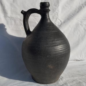 May include: A dark, matte black ceramic jug with a handle and spout. The jug has a rounded body and a textured surface. The handle is arched, and the spout is small and cylindrical. The jug is likely used for holding liquids.
