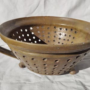 May include: A brown ceramic colander with a handle and small feet. The colander has numerous small holes. It is designed for draining and rinsing food. The colander is on a white surface.