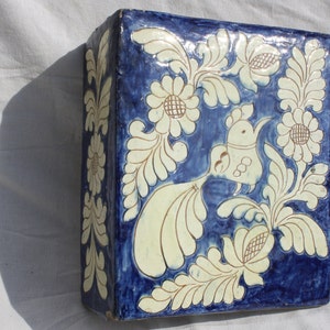 Op de afbeelding: Een vierkante keramische tegel met een blauwe achtergrond en een wit bloemmotief. De tegel heeft een gestileerde vogel met een lange staart en een bloemmotief.