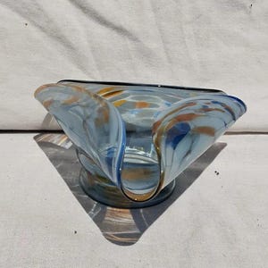 Puede incluir: Un cuenco decorativo de vidrio con una forma asimétrica única. El cuenco presenta un color base azul claro con patrones arremolinados de naranja y azul oscuro. El borde está delineado con un borde negro. El cuenco se asienta sobre una pequeña base circular.