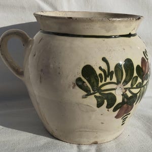 Puede incluir: Una jarra de cerámica color crema con asa y boca ancha. La jarra está decorada con diseños florales verdes y granates. Una banda verde oscuro rodea la parte superior. La jarra es probablemente vintage o antigua.