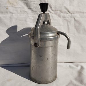 May include: A vintage, cylindrical, aluminum espresso maker with a black handle and a curved spout. The handle is arched, and the top has a black knob. The espresso maker is sitting on a white surface.