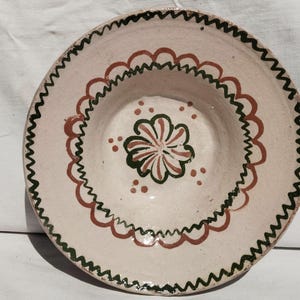 May include: A round ceramic bowl with a cream-colored base featuring hand-painted decorations. The design includes a dark green zigzag border along the rim, a scalloped terracotta-colored ring, and a central flower motif in green and terracotta.