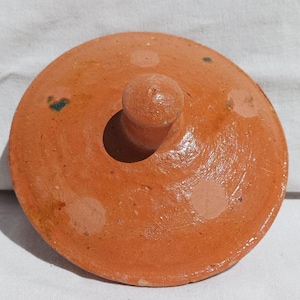 Op de afbeelding: Een terracotta deksel met een knop aan de bovenkant. Het deksel is versierd met witte en groene stippen.