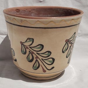 Può includere: Un vaso di ceramica beige con bordo marrone e un motivo decorativo a foglie verdi, nere e rosse. Il vaso ha una forma leggermente svasata e un design semplice, adatto per uso interno o esterno.