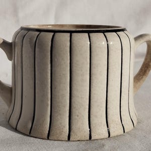 May include: Cream-colored ceramic sugar bowl with two handles and vertical black stripes. The rim and the top edge of the bowl are outlined in black. The sugar bowl has a rounded shape and is likely vintage.