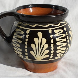 May include: A handmade ceramic mug with a black exterior, adorned with cream-colored floral and geometric patterns. The interior and base are a warm orange-brown. Features a curved handle for easy gripping.