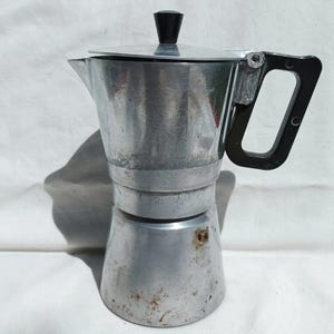 May include: A vintage aluminum stovetop espresso maker with a black handle and lid knob. The coffee pot has a classic design, showing signs of use and age. It is a traditional Italian coffee maker.