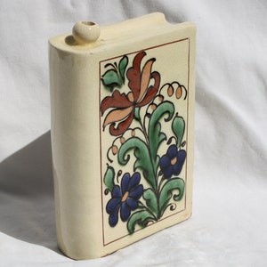 May include: A ceramic book-shaped vase with a floral design. The vase is painted with green leaves, orange and red flowers, and blue flowers. The vase has a cream background.
