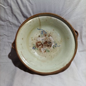 May include: A large, round, ceramic bowl with two handles. The bowl is decorated with a floral design in shades of blue, green, and brown. The bowl has a crack on the inside.