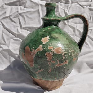 May include: A green ceramic jug with a handle and a rounded body. The jug has a distressed appearance with brown patches and a light brown base. The handle is curved, and the jug has a spout at the top.