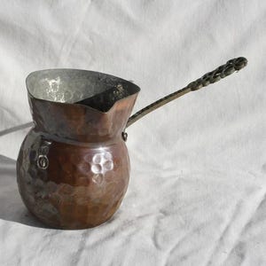 Puede incluir: Una cafetera de cobre martillado con un asa decorativa. La cafetera tiene un cuerpo redondeado y un pico.