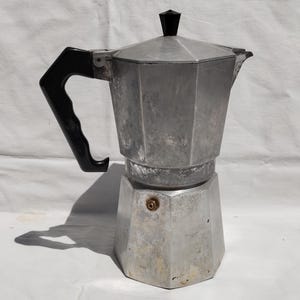 May include: A silver stovetop espresso maker with a black handle and lid knob. The coffee pot has a faceted design and is sitting on a white surface. The espresso maker is a kitchen appliance.