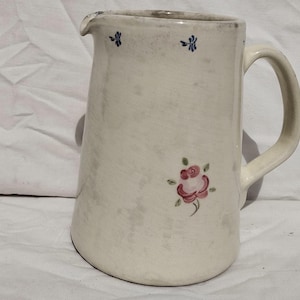 May include: A cream-colored ceramic pitcher featuring a hand-painted pink rose with green leaves on the side and small blue floral accents near the rim. The pitcher has a curved handle and a pouring spout, with a smooth, slightly mottled finish.