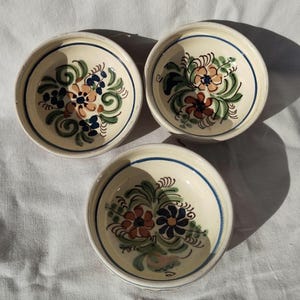 May include: Three small, round ceramic bowls with floral designs. Each bowl features a cream-colored base, a blue rim, and a hand-painted floral pattern in shades of brown, blue, and green. The bowls are arranged on a white surface.
