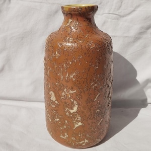 Puede incluir: Un jarrón alto de cerámica marrón con una superficie texturizada y un cuello estrecho. El jarrón tiene un patrón moteado con acentos de color más claro. El borde del jarrón es de color amarillo claro. Es un objeto decorativo.