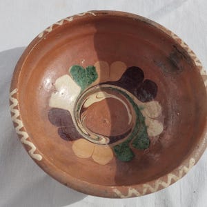 Op de afbeelding: Een handgemaakte terracotta-kleurige keramische kom met een decoratief bloemmotief in het midden. Het ontwerp bevat groene, witte, paarse en bruine bloemblaadjes. De rand heeft een wit zigzagpatroon.