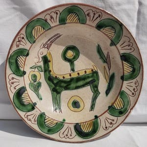 Puede incluir: Un plato redondo de cer&aacute;mica con una base de color crema moteado que presenta una gacela verde pintada a mano en el centro. El borde est&aacute; decorado con un patr&oacute;n repetitivo de arcos verdes que contienen l&iacute;neas verticales amarillas y motivos florales marrones.