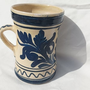 Könnte beinhalten: Eine weiße Keramiktasse mit blauem Blumendesign und blauem Rand. Die Tasse hat einen Henkel und eine wellenförmige blaue Linie am Boden.