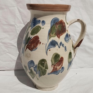 Może przedstawiać: Ceramiczny dzbanek o kremowej podstawie z ręcznie malowanymi abstrakcyjnymi wzorami kwiatowymi w odcieniach niebieskiego, zielonego i czerwonobrązowego. Naczynie ma szeroki, zaokrąglony korpus, pojedynczy zakrzywiony uchwyt i brązowy brzeg.