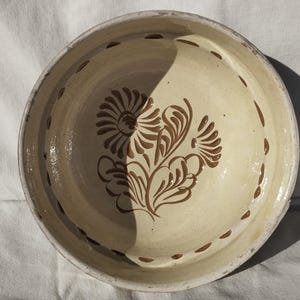 May include: Cream-colored ceramic bowl with a floral design in brown. The bowl has a wide rim with small, evenly spaced brown dots. The floral design features stylized flowers and leaves. The bowl is likely handmade.