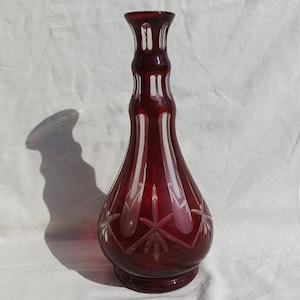Könnte beinhalten: Eine rubinrote Glaskaraffe mit breiter Basis und langem, schlanken Hals. Die Karaffe ist mit aufwendigen Schliffmustern verziert.