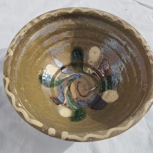 Pode incluir: Uma tigela de cerâmica feita à mão com uma estética rústica e terrosa. A tigela apresenta um exterior castanho claro com uma borda ondulada de cor creme. O interior tem um design em espiral em tons de verde, azul e castanho.
